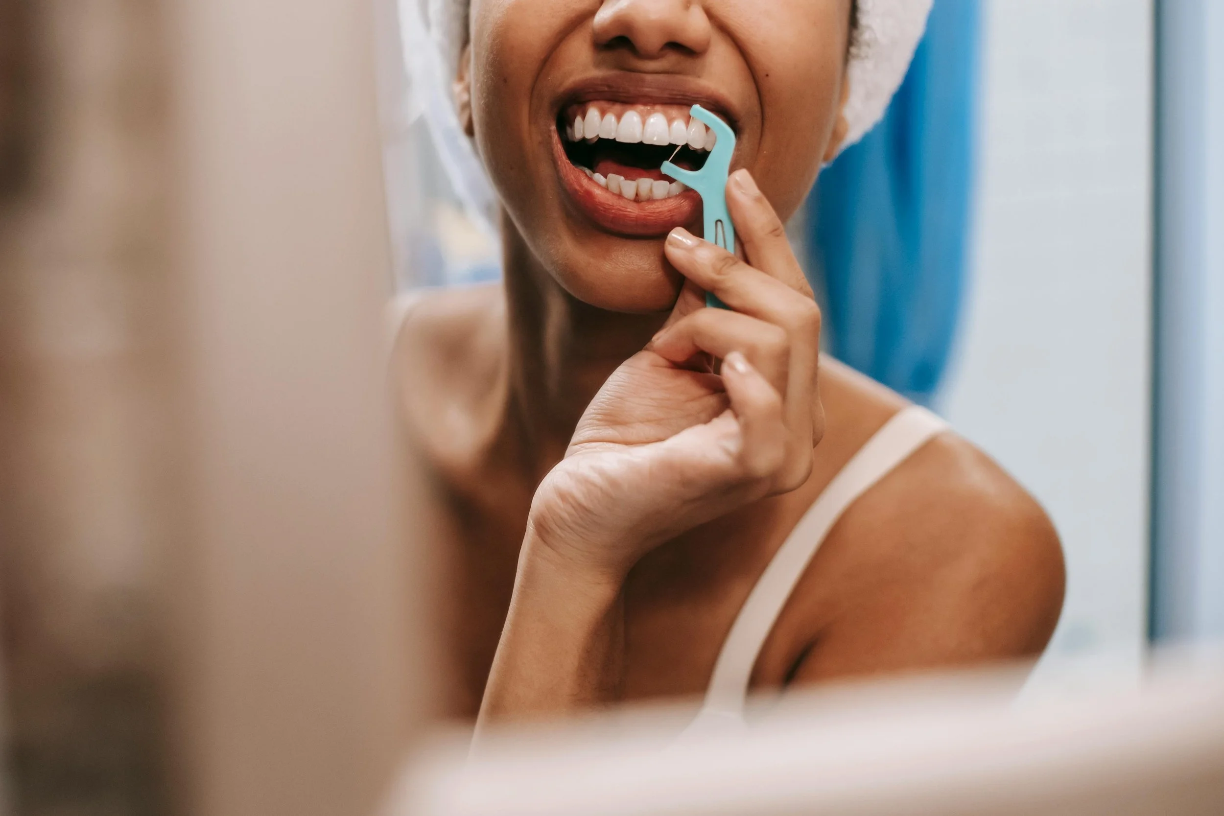 Woman flossing her teeth with a dental pick while smiling in front of a mirror.