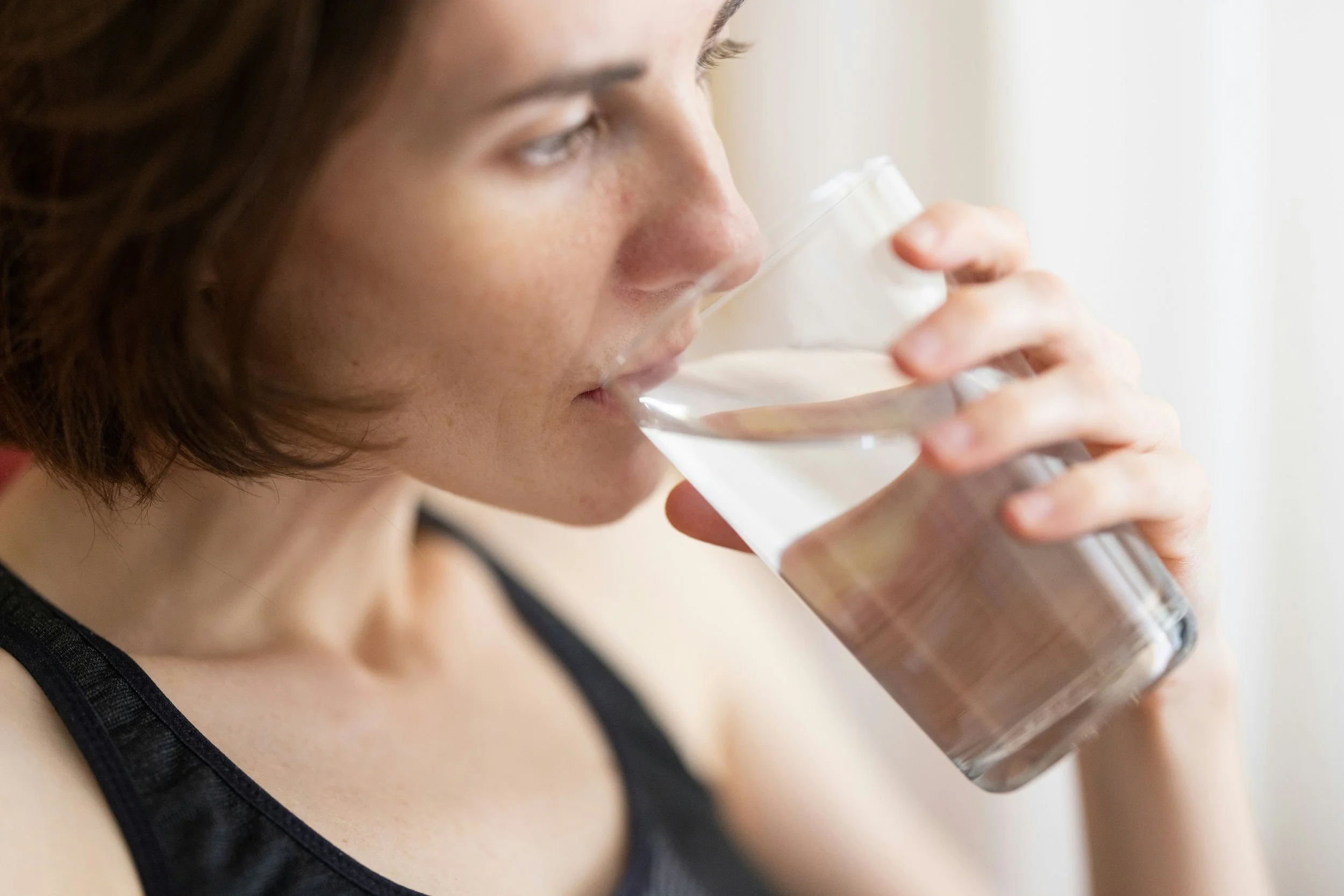 Woman drinking a glass of water, viewed from the side, wearing a black tank top and appearing to hydrate in a calm indoor setting.