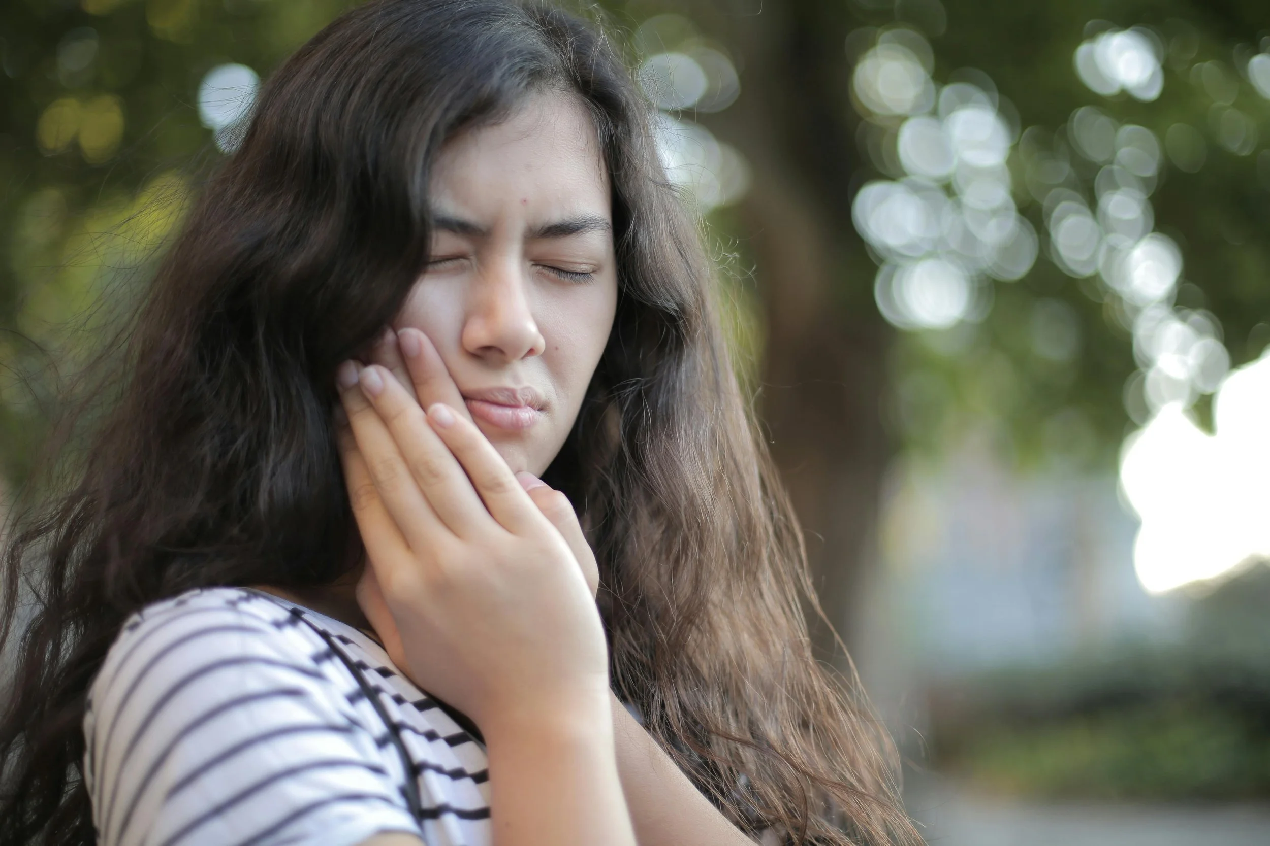 Young woman outdoors holding her cheek in pain, showing signs of tooth sensitivity