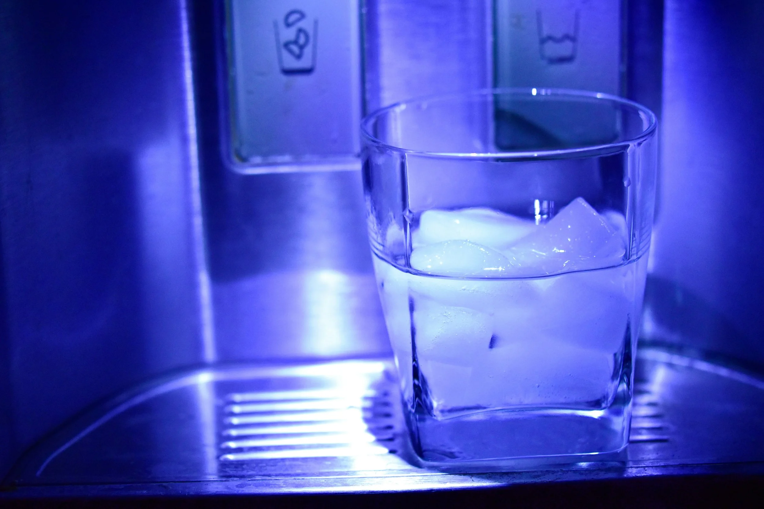 Glass of ice water placed under a refrigerator water and ice dispenser illuminated with blue lighting.