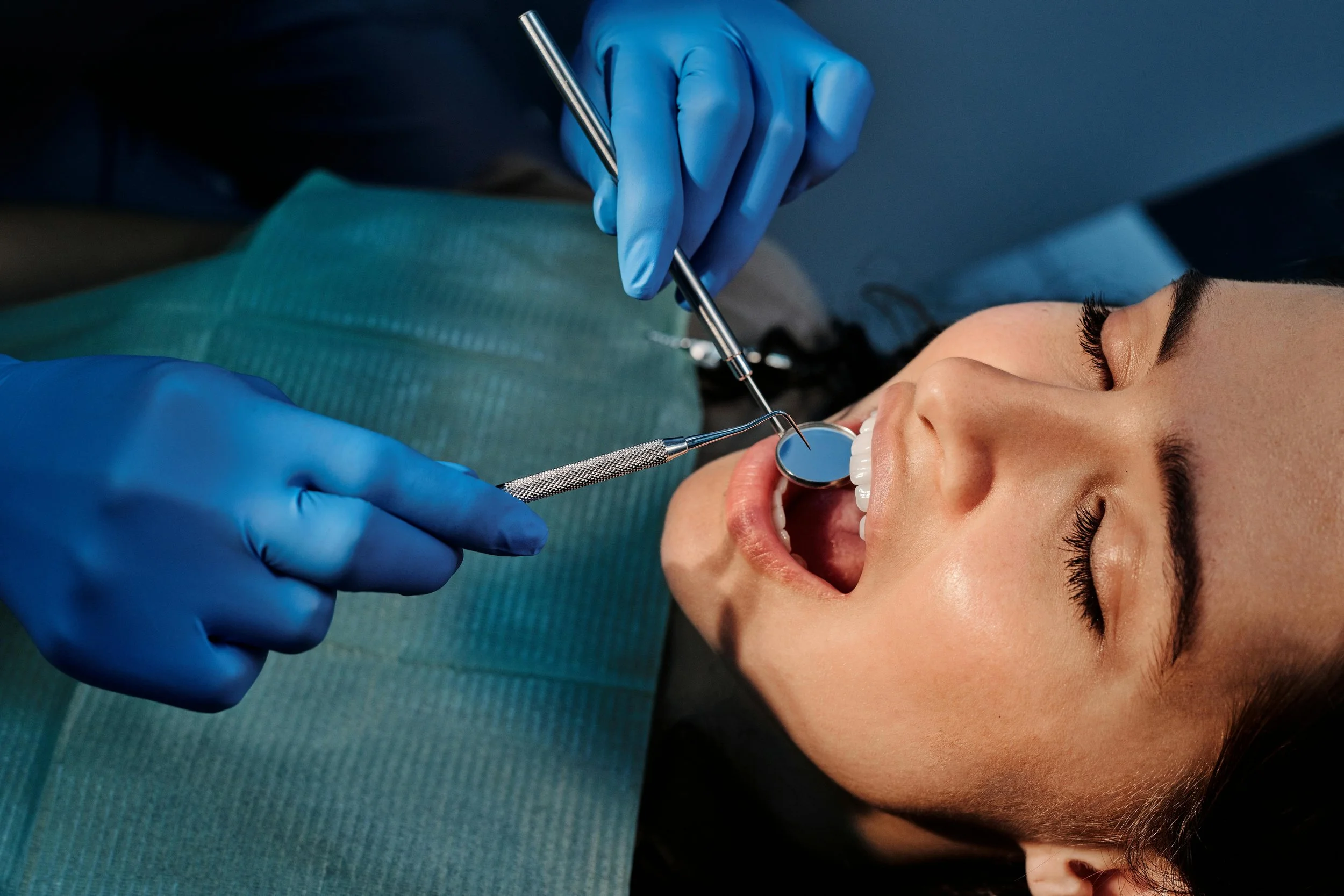 Dentist wearing blue gloves examining a woman's teeth with dental tools during a checkup.