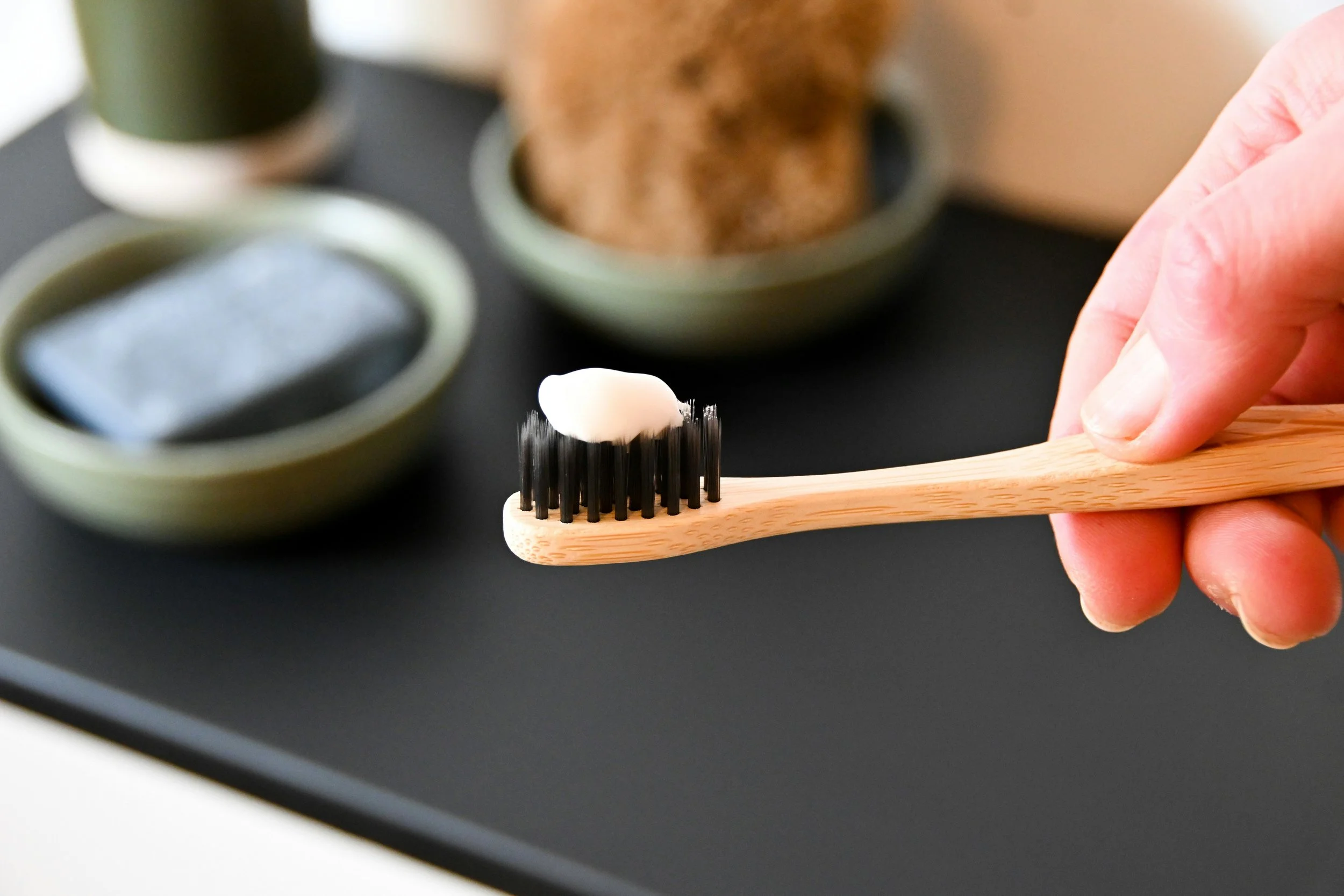 Close-up of a hand holding a bamboo toothbrush with black bristles and a small amount of toothpaste, set against a bathroom counter background.