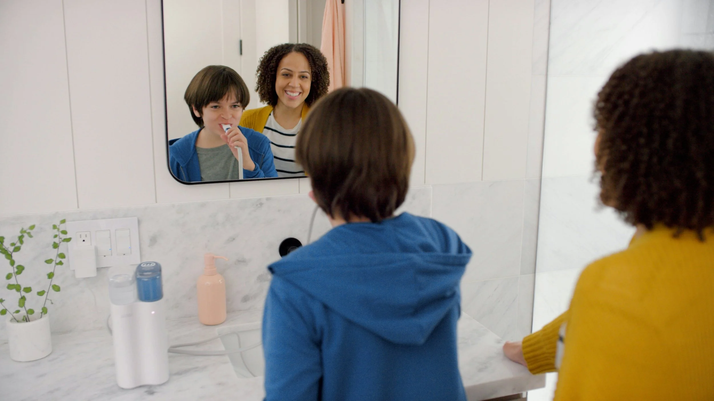 Child brushing their teeth at a bathroom sink while a smiling adult stands beside them, both reflected in the mirror above the counter.