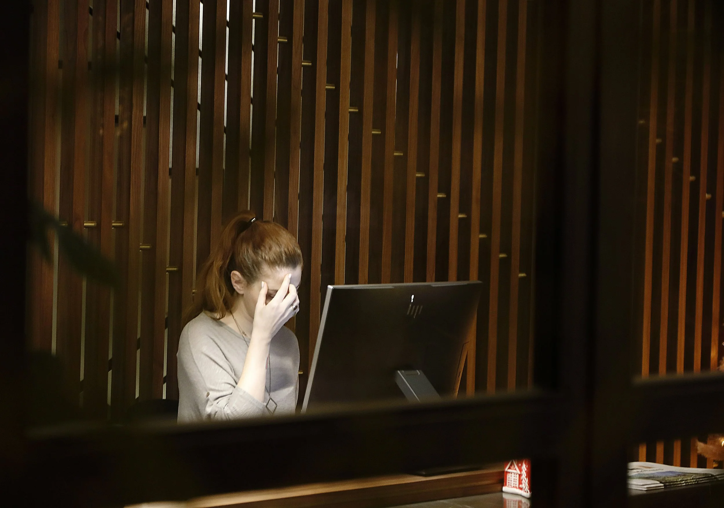 Woman sitting at a desk in front of a computer with her hand on her forehead, appearing stressed or fatigued, in a dimly lit office with wooden paneling.