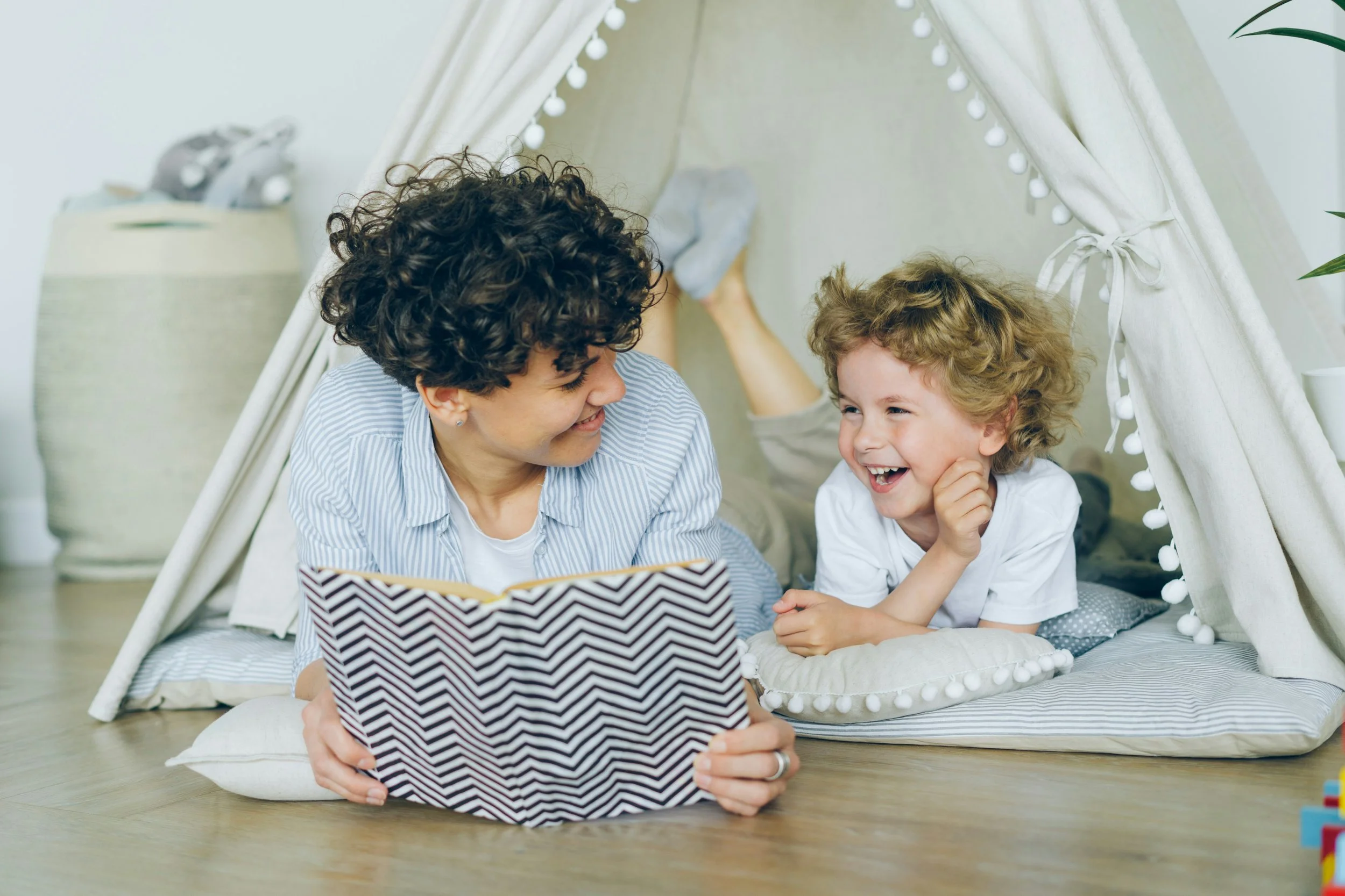 A smiling adult and young child lying on the floor inside a cozy fabric tent, sharing a moment while reading a book together in a playful indoor setting.