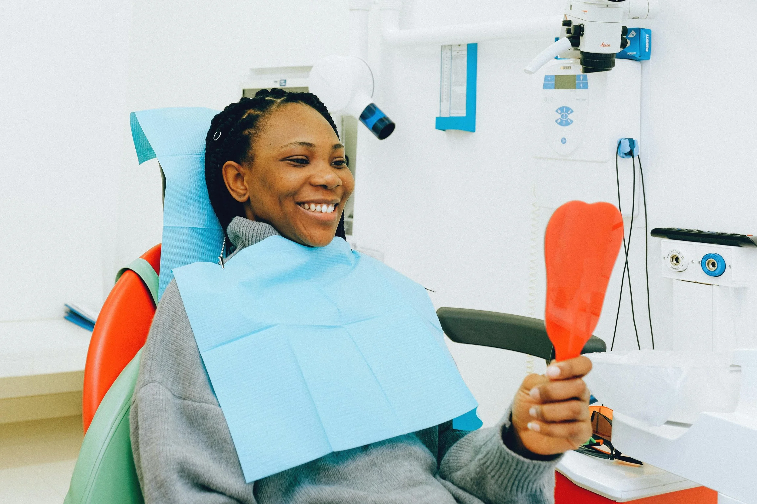 Smiling woman sitting in a dental chair holding a red mirror and looking at her teeth after a dental appointment.
