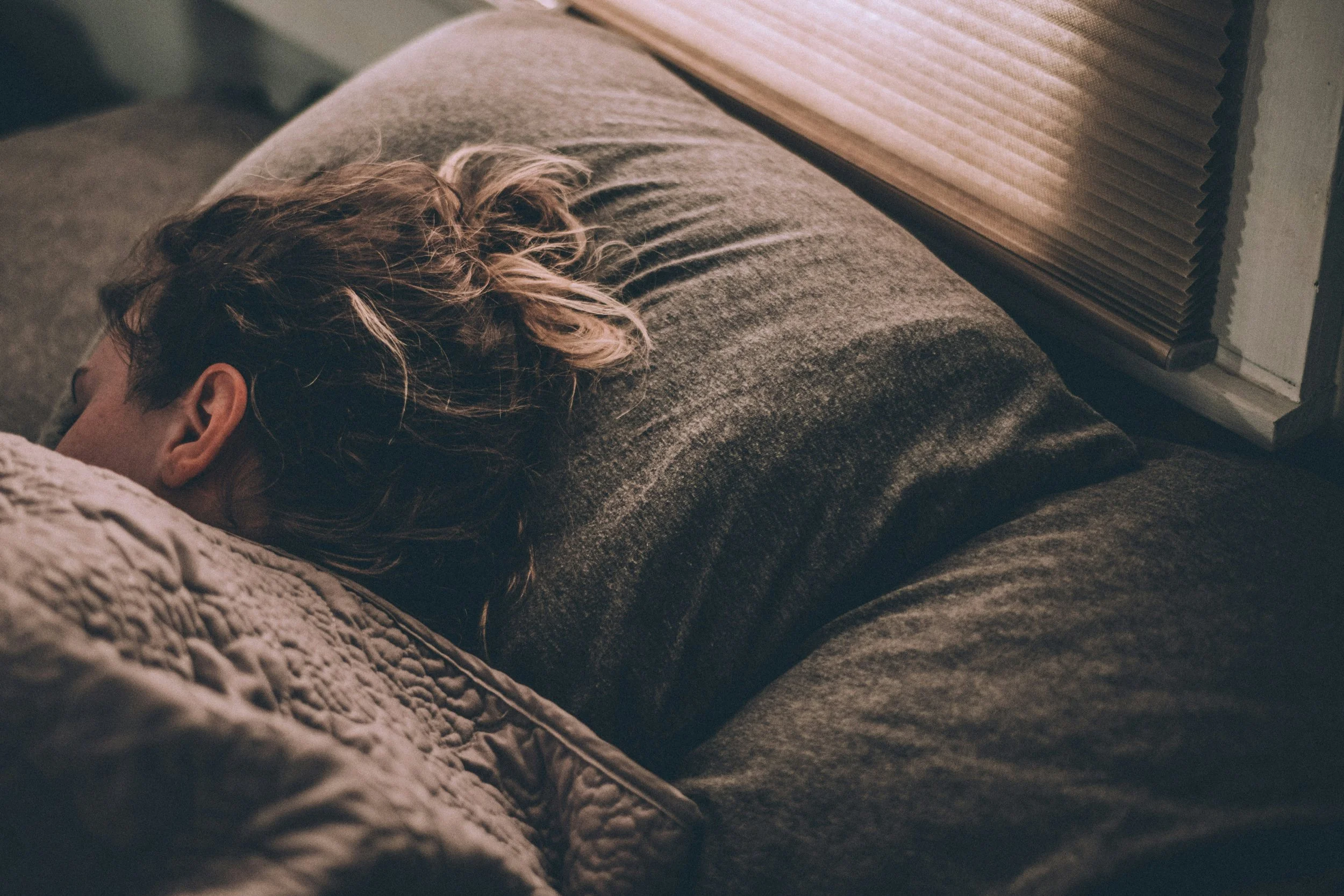 Person sleeping peacefully in bed with their head resting on a dark pillow and wrapped in a quilted blanket near a window with closed blinds.