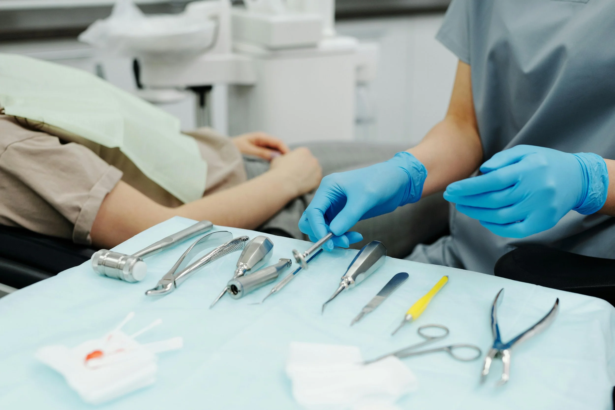 Dentist wearing blue gloves preparing dental instruments on a tray next to a patient in a dental chair.
