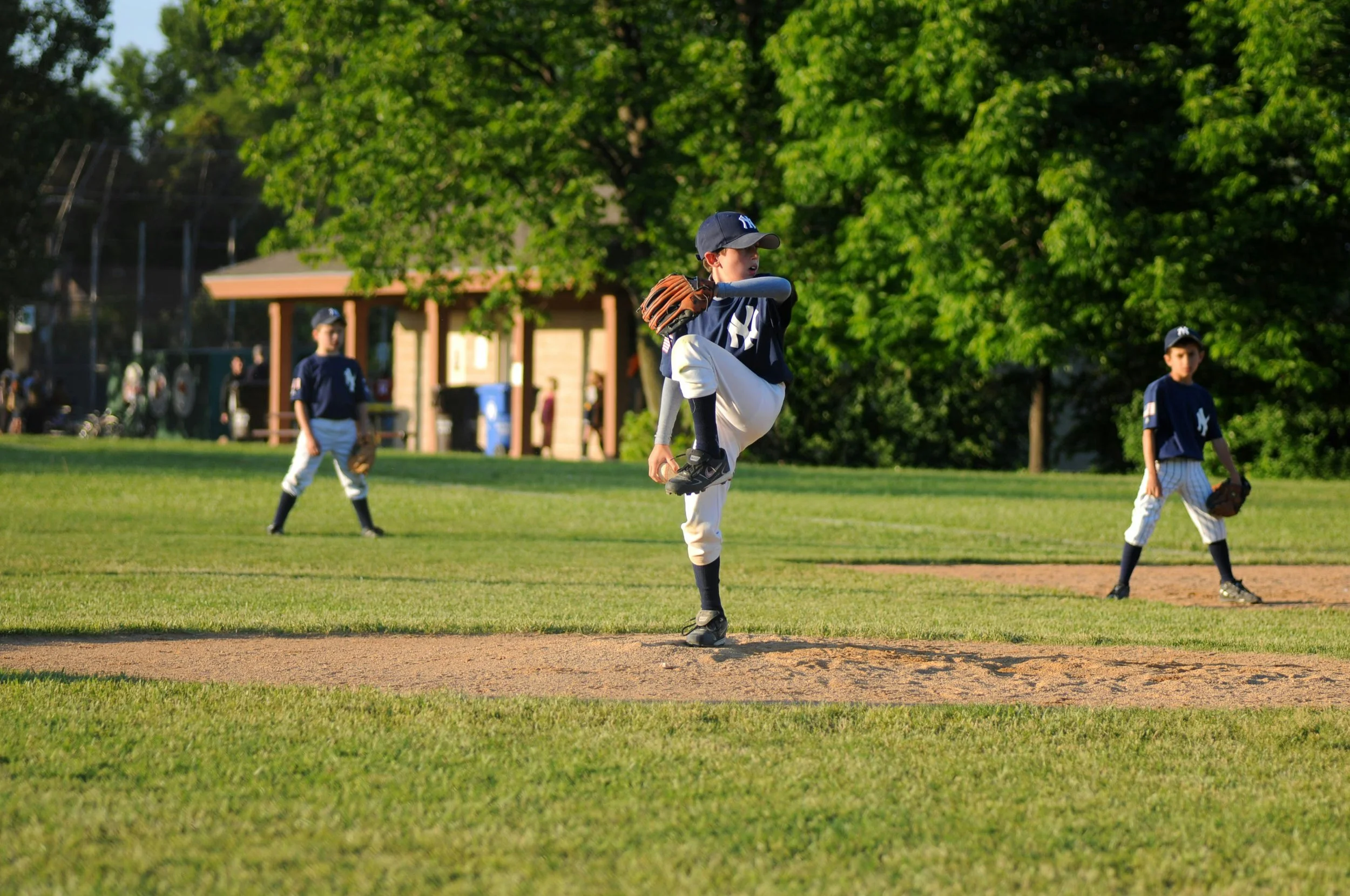 Youth baseball pitcher mid-windup on the mound with teammates in the field during a sunny afternoon game at a local park.