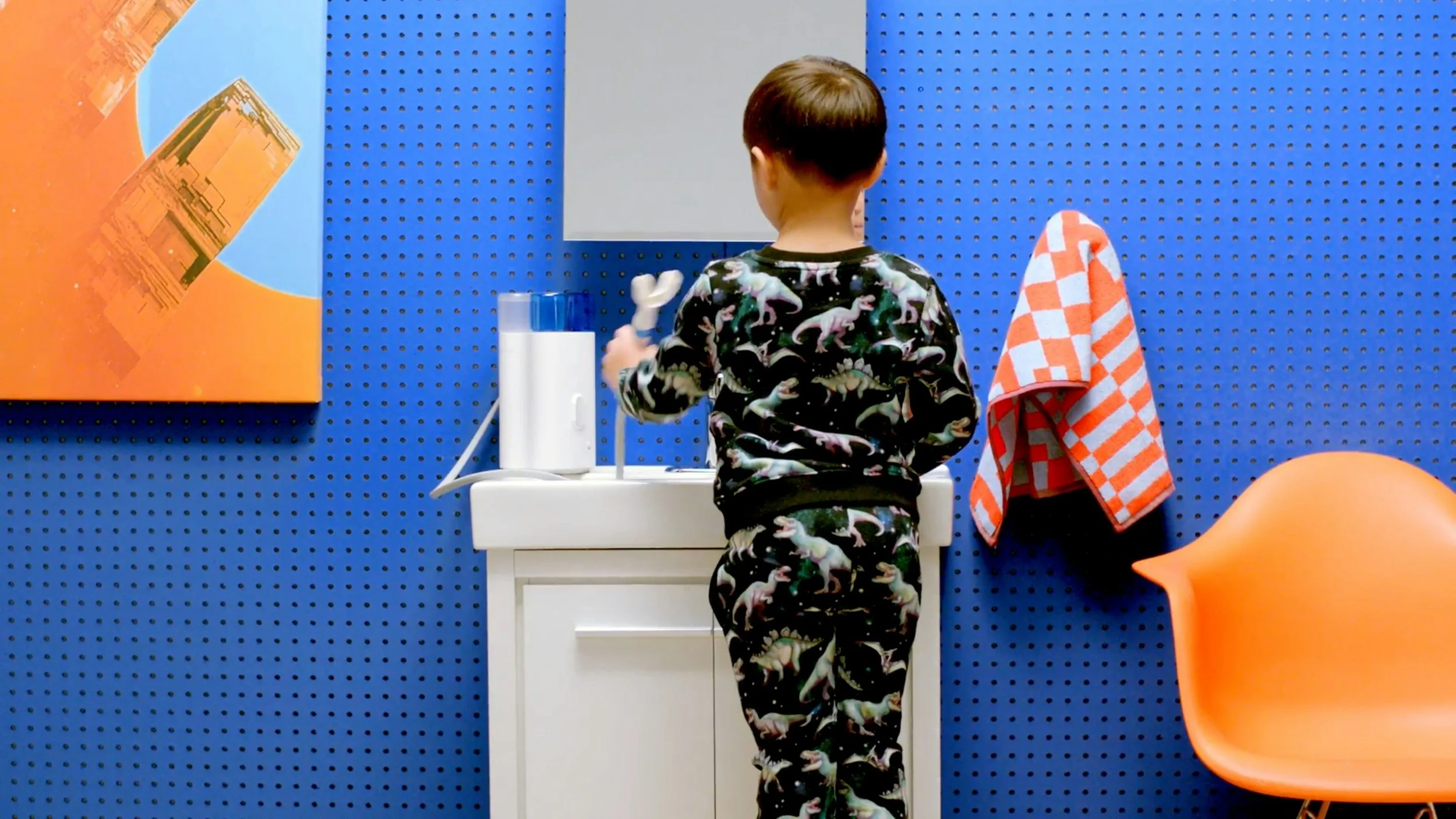 Young child in dinosaur pajamas standing at a bathroom sink holding a toothbrush, with a blue pegboard wall, orange chair, and checkered towel in the background.